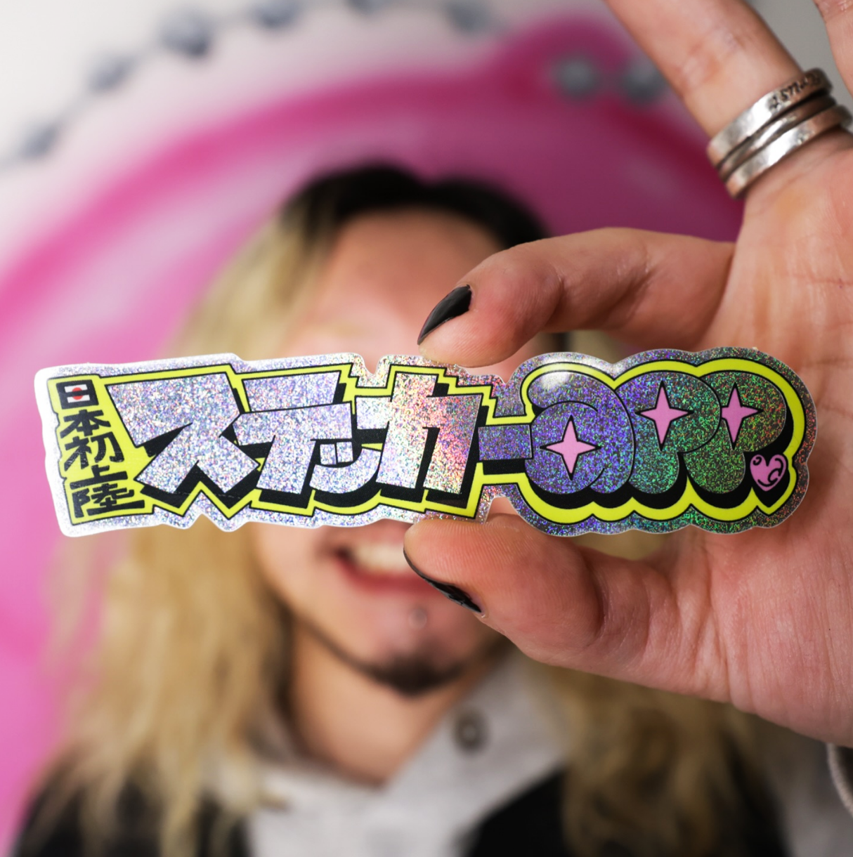 Person holding a colorful, holographic Japanese sticker with playful, stylized text and designs, smiling in the blurred background.