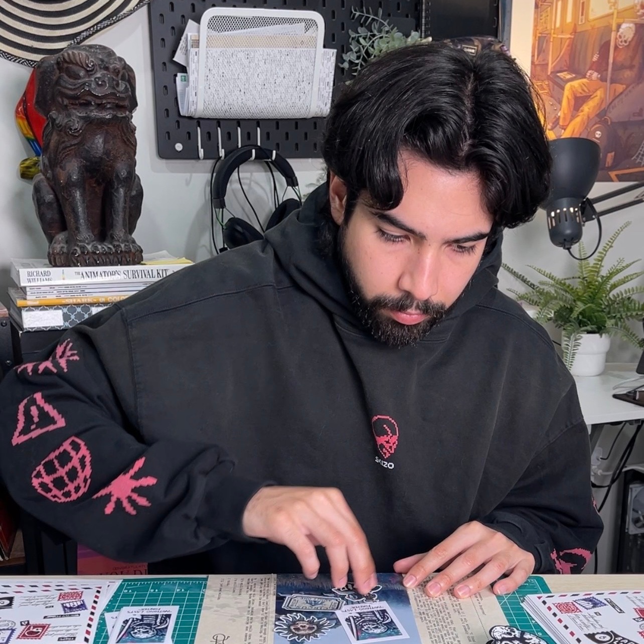 Een man in een zwarte hoodie zit in zijn atelier en legt stickers neer op de tafel voor hem, met beelden en kunst op de achtergrond.