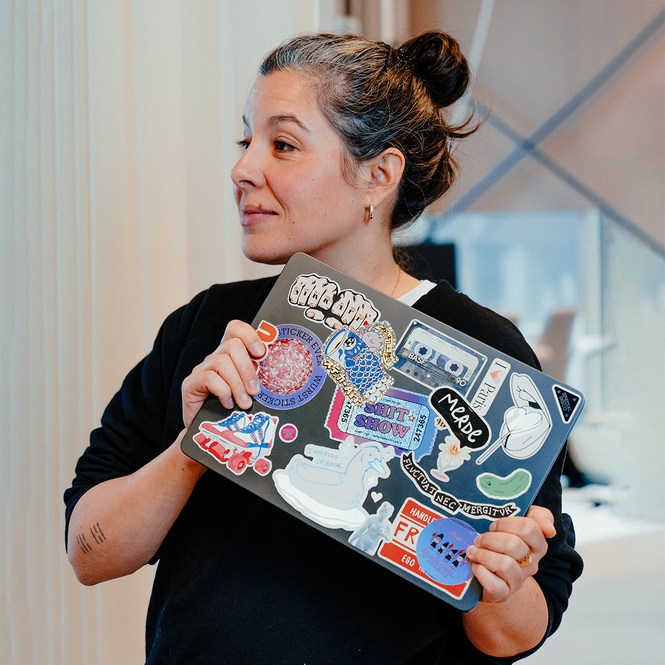 Person holding a laptop covered in colorful stickers, including illustrations, logos, and graphic designs, in a bright indoor setting.