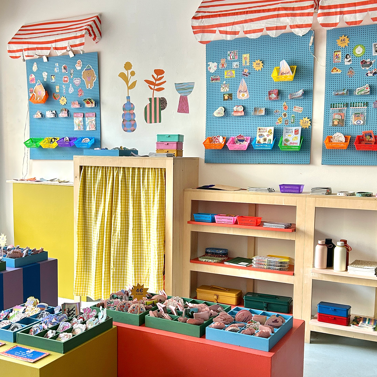 A colorful craft shop with pinboards displaying artsy stickers, shelves showcasing various stickers, and decorative red-and-white striped awnings.