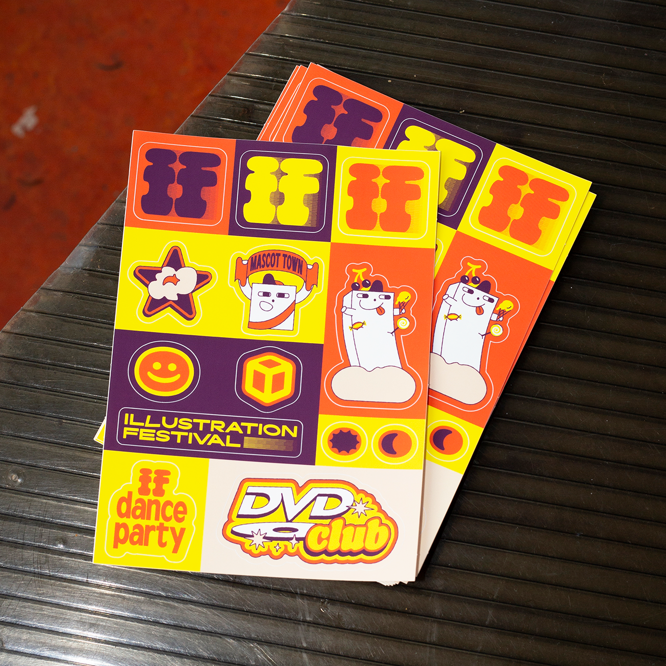 Two sheets of colorful stickers on a surface, featuring cheerful illustrations, text like "Dance Party" and "DVD Club," and a cartoon character.