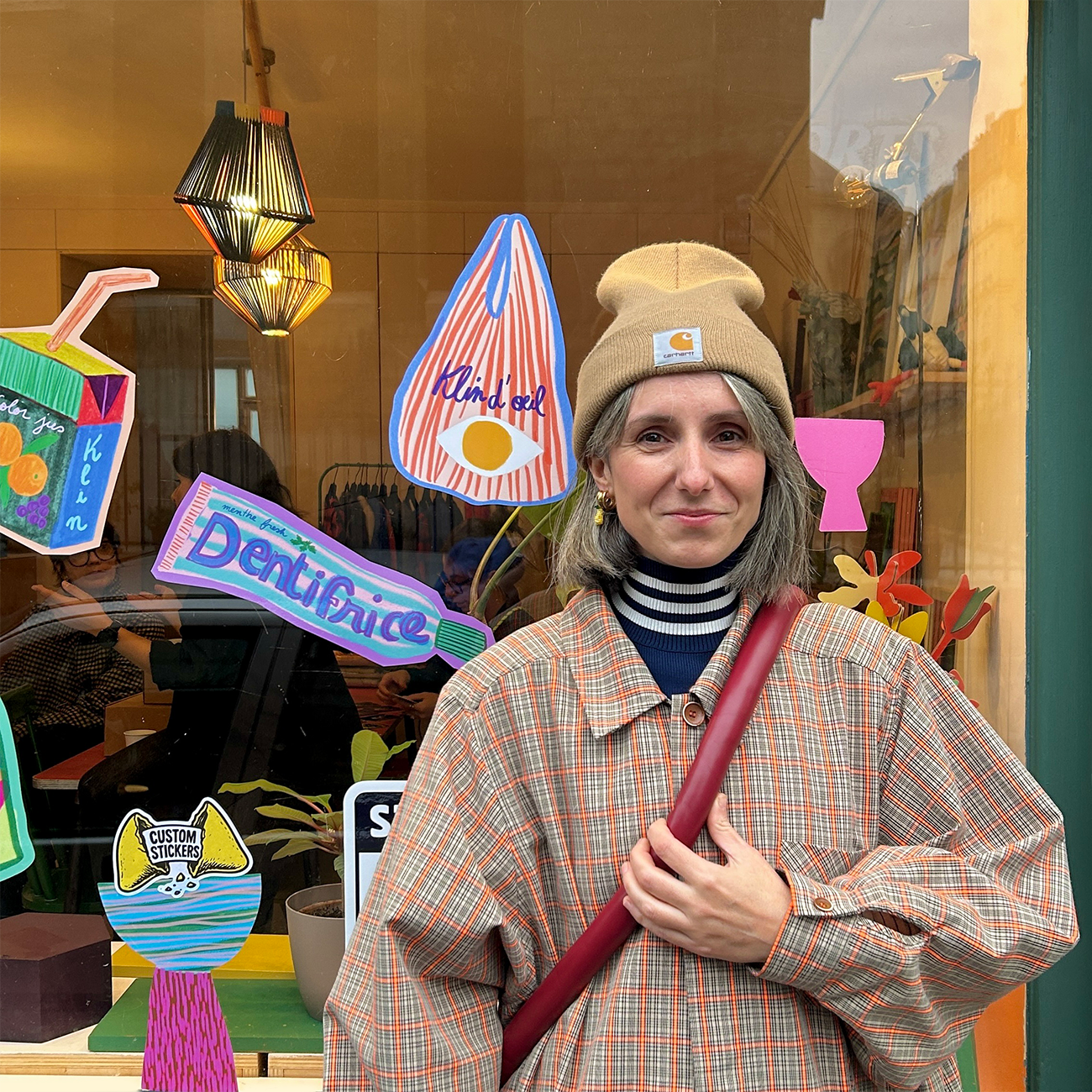 Person in plaid coat and beanie stands by a colorful shop window display featuring playful, oversized product cutouts.