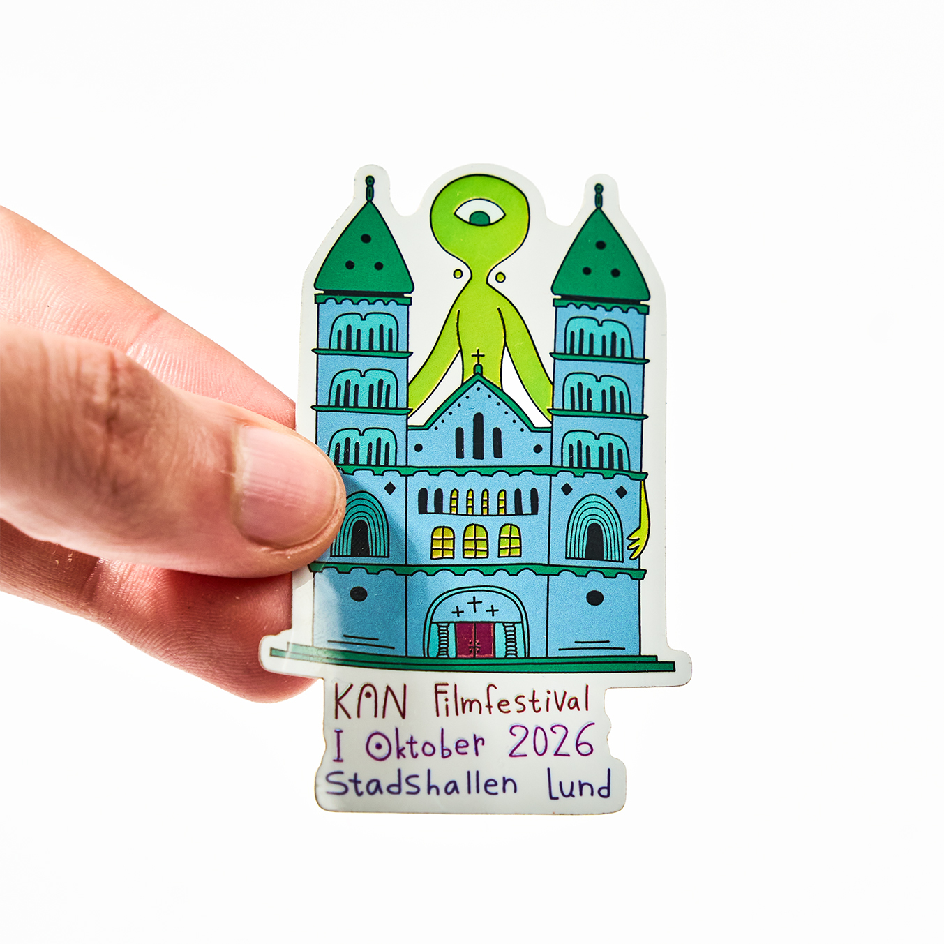 Hand met een contourgesneden sticker voor KAN Filmfestival, oktober 2026 in Stadshallen Lund, met een kathedraal en een groen eenogig buitenaards wezen