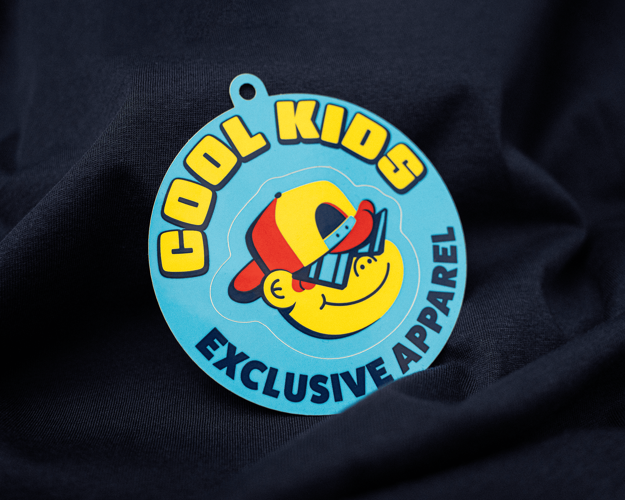 A circular hang tag on fabric with a smiling face wearing a cap and sunglasses, labeled "Cool Kids Exclusive Apparel" in bold letters.