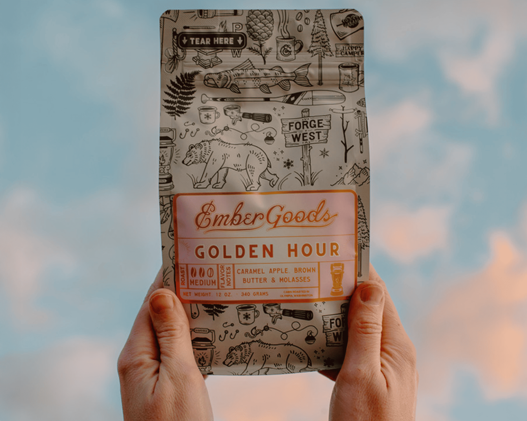 Handen met een zak Golden Hour-koffie van Ember Goods met een metallic etiket tegen een achtergrond van lucht