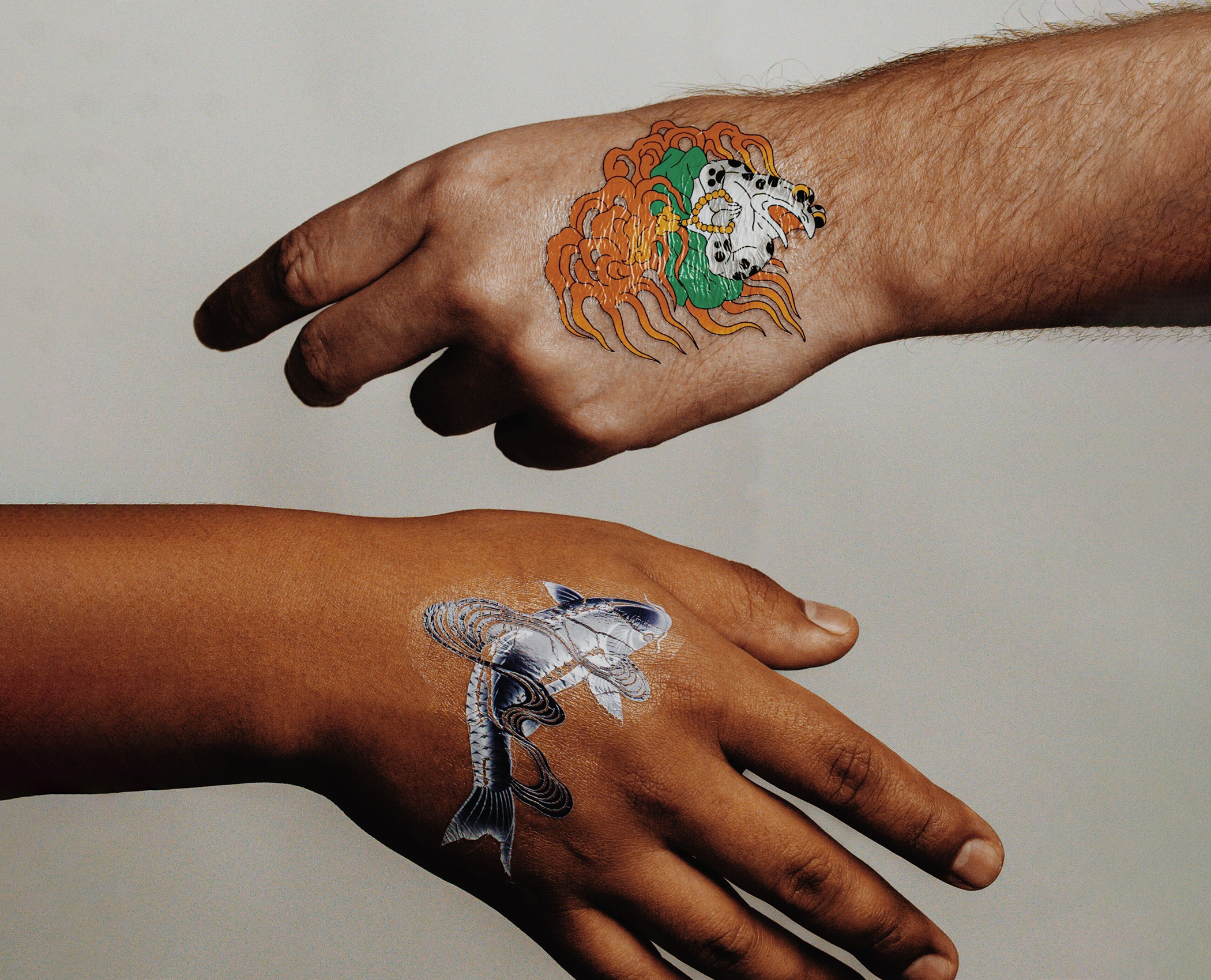 Temporary tattoos on two hands, one featuring a detailed koi fish design and the other a stylized skull with flowing orange elements, set against a neutral background.