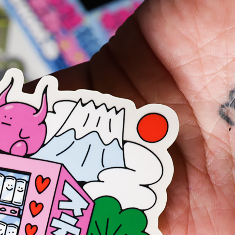 Hand holding a sticker with a pink cartoon character, mountain, and clouds, surrounded by colorful shapes.
