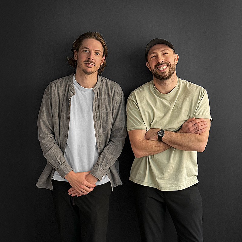 Two men standing in front of a dark wall, one wearing a gray shirt over a white tee, the other in a light tee and cap, both smiling.